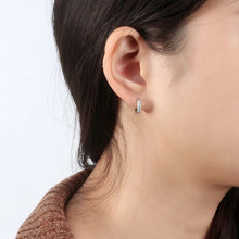 A close-up of a model's ear wearing a small silver huggie hoop earring with clear stones. The brand logo 'Orsa Jewels' is visible in the top-left corner.