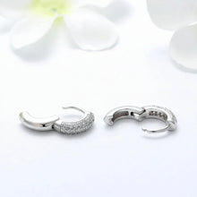 A pair of silver-toned huggie hoop earrings with pavé-set clear stones on one side, displayed on a clean white background with soft white flower petals. The Orsa Jewels logo is visible in the top-left corner.