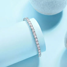Silver bracelet with clear stones on a light blue background, featuring a brand logo.

