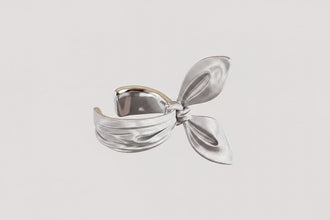 Silver bow-shaped ring on a light gray background with 'GEEZENCA' branding.