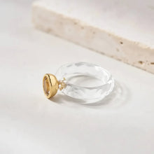 Clear ring with gold base on a light wooden surface, branded 'GEEZENCA'.