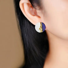 Close-up of an ear wearing a round earring with gold and purple design, brand 'GEEZENCA' visible.