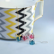 Three colorful necklaces displayed in front of a mug with a zigzag pattern, branded 'Geezenca'.