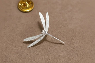 Silver dragonfly-shaped brooch on a brown background with 'GEEZENCA' branding.