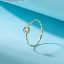 A gold ring with a single round clear gemstone in a bezel setting, displayed on a light blue background with a soft shadow. The brand logo 'Orua Jewels' is visible in the top-left corner.