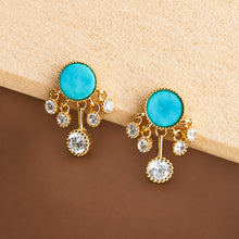 Gold earrings with turquoise stones and small crystals on a beige surface, branded 'Geezenca'.