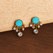 Gold earrings with turquoise stones on a textured surface, featuring the brand 'Geezenca'.