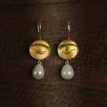 Gold earrings with pearl drops on a dark textured background, featuring the brand 'Geezenca'.