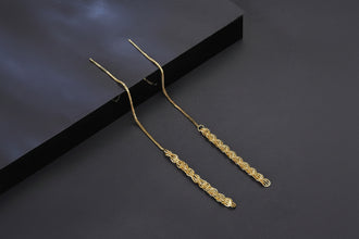 A pair of gold-toned long dangle earrings with a delicate chain and an ornate braided drop, displayed on a dark gray surface with a black box in the background. The brand logo 'Orusa Jewels' is visible in the top-left corner.