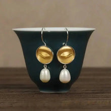 Gold earrings with pearl drops on a dark green cup against a brown background, featuring the brand 'Geezenca'.