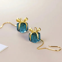 Teal gift box earrings with gold ribbons on a beige background, featuring the brand 'Geezenca'.