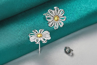Flower-shaped earrings with gold centers on a teal fabric background, featuring the brand 'Doteffal'.