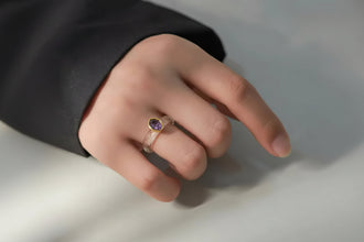 Hand wearing a ring with a purple gemstone on a neutral background, featuring the brand 