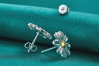 Silver flower-shaped earrings with a gold center on a teal fabric background, featuring the brand 'DOKEFFIL'.