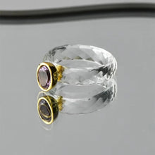 Clear gemstone ring with gold and purple accents on a reflective surface, 
