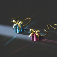 Earrings shaped like gift boxes with gold ribbons on a dark background, branded 'Geezenca'.