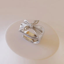 Silver bow-shaped ring on a light beige background with 'GEEZENCA' branding.
