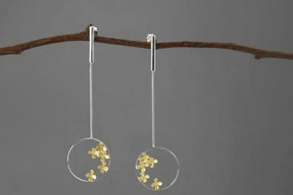 Pair of earrings with floral design in clear circles on a branch against a gray background, branded 'Geezenca'.