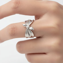 Silver ring with a bow design worn on a finger, brand 'Geezenca' visible.