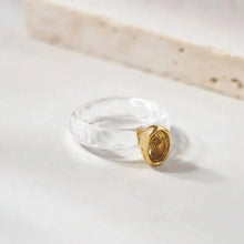 Gold ring with a clear stone on a light background, branded 'Geezenca'.