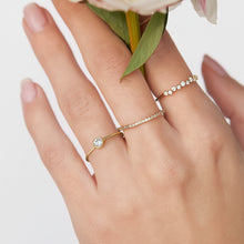 Three delicate gold rings with clear gemstones worn on a hand, with a soft pink flower stem in the background. The brand logo 'Orna Jewels' is visible in the top-left corner.