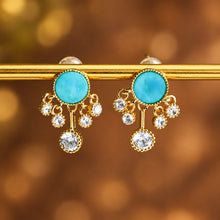 Gold earrings with turquoise stones and small crystals on a blurred background, featuring the brand 'Geezenca'.